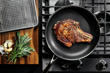 A large steak being seared in a pan, and a cutting board with herbs on top next to it.