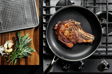 A large steak being seared in a pan, and a cutting board with herbs on top next to it.
