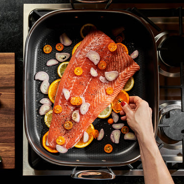 A large salmon filet being seasoned and covered in vegetables and and citrus fruit slices, sitting inside the HexClad Hybrid Roasting Pan, shown from a bird