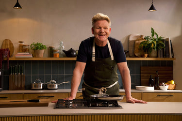 Chef Gordon Ramsay standing behind a kitchen counter wearing an apron, smiling for the camera.