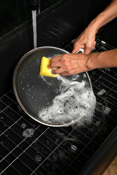 A person scrubbing a HexClad pan under running water with a soapy yellow sponge.