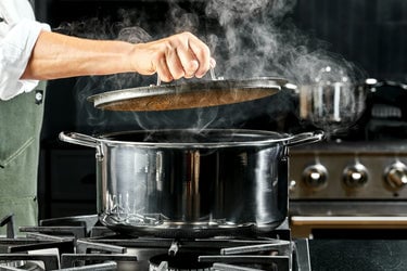 A hand taking the lid off of a steaming, stainless steel pot.