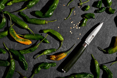 Many shishito peppers on a dark grey surface, next to a HexClad knife.