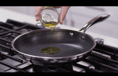A hand pouring a ramekin of oil into a HexClad hybrid frying pan.