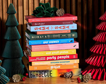 A stack of cookbooks with vibrant covers, surrounded by decorative pine cones and holiday trees.