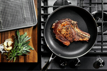A large steak in a HexClad frying pan, shown from a bird’s-eye view, with herbs and garlic gloves on the counter next to it.