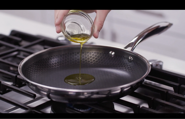 A person pouring a small ramekin of cooking oil on to the surface of a HexClad pan.
