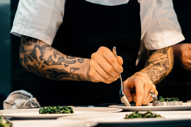 A person with tattoos on both arms is plating several dishes with a green salad, using a spoon and a food ring.