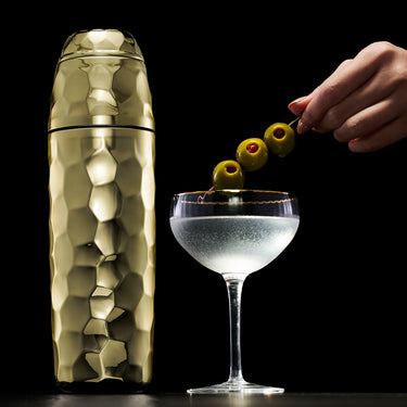 Gold hexclad cocktail shaker next to a cocktail made with olives on a black background