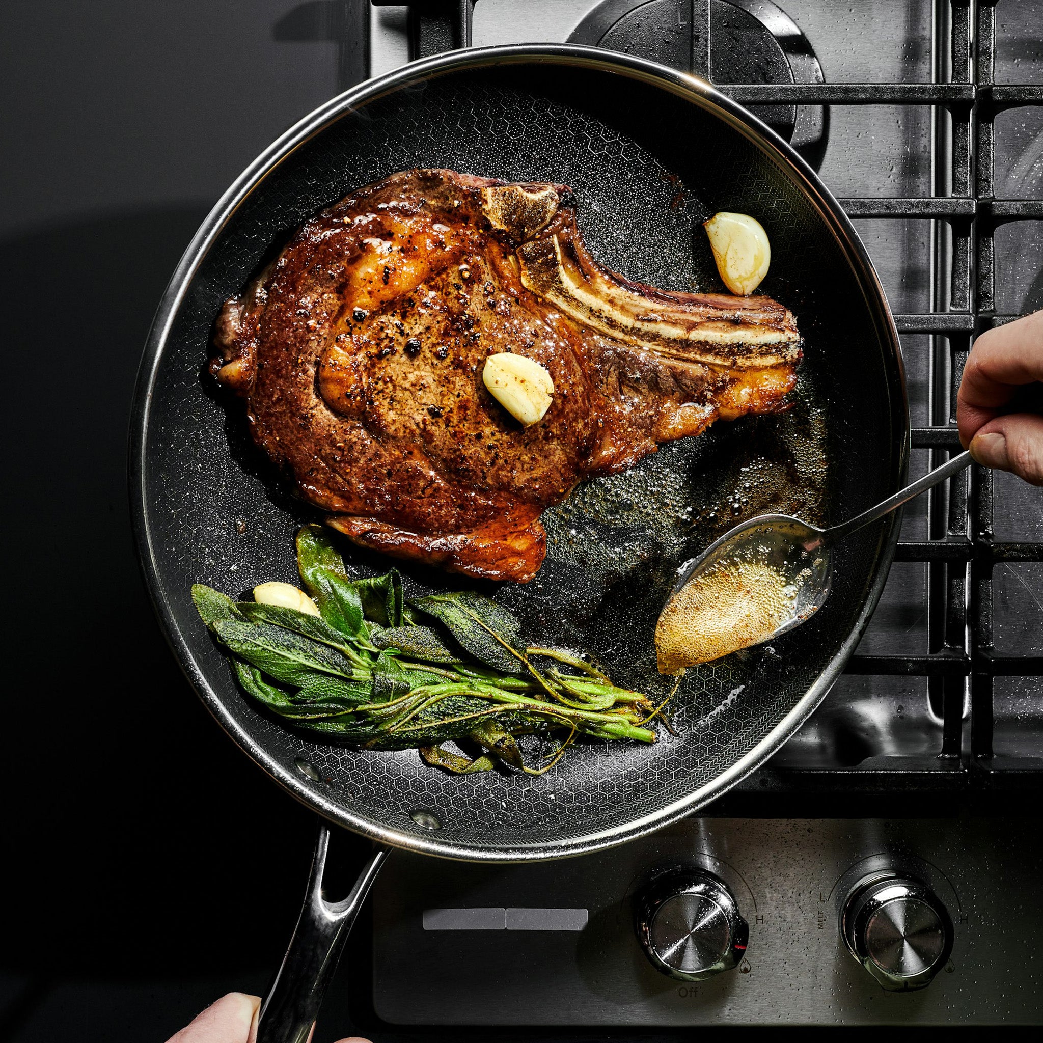 A ribeye steak cooking in a pan with garlic and herbs on a stovetop.
