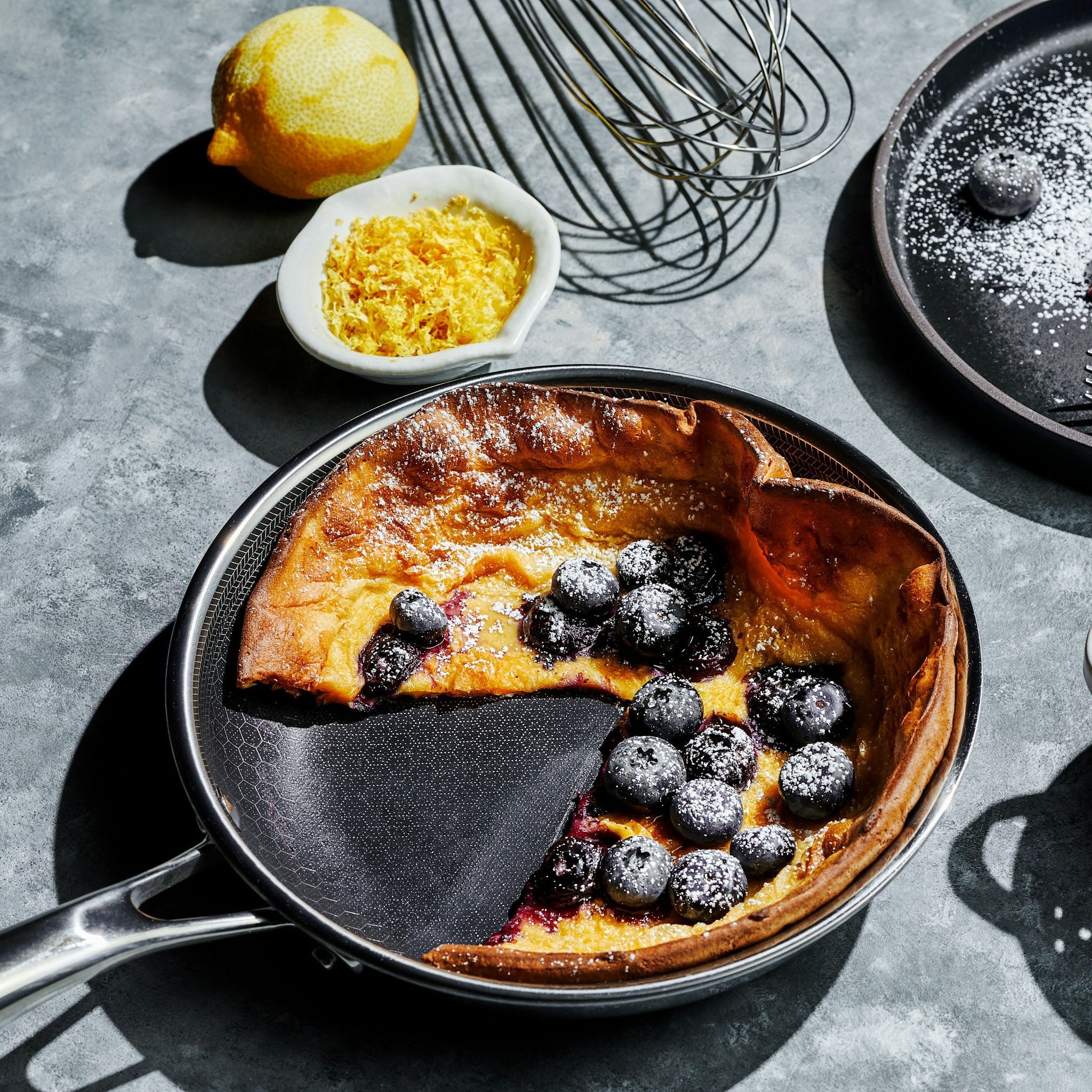 Dutch baby pancake with blueberries, lemon zest, and a whisk on a gray surface.