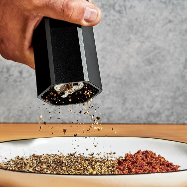 Hand using a black pepper grinder over a plate with spices.