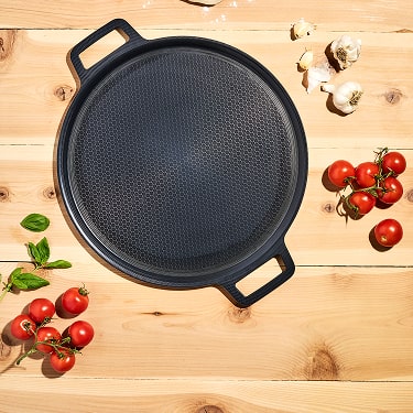 Black round pan on wooden surface with tomatoes, garlic, and basil.