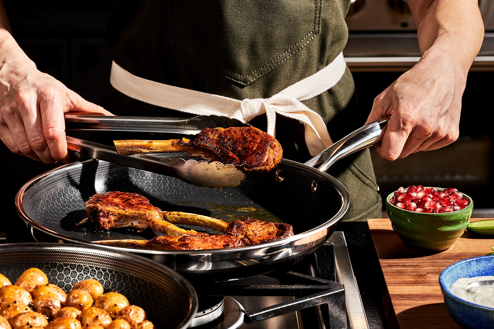 Person cooking lamb chops in a pan with pomegranate seeds nearby.