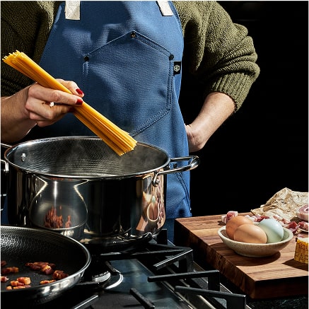 Person in blue apron cooking pasta on a stove with ingredients nearby.