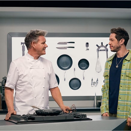 Two people talking in a kitchen with cooking utensils on the wall.