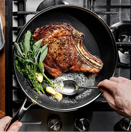 Person cooking a steak with herbs and garlic in a pan on a stove.