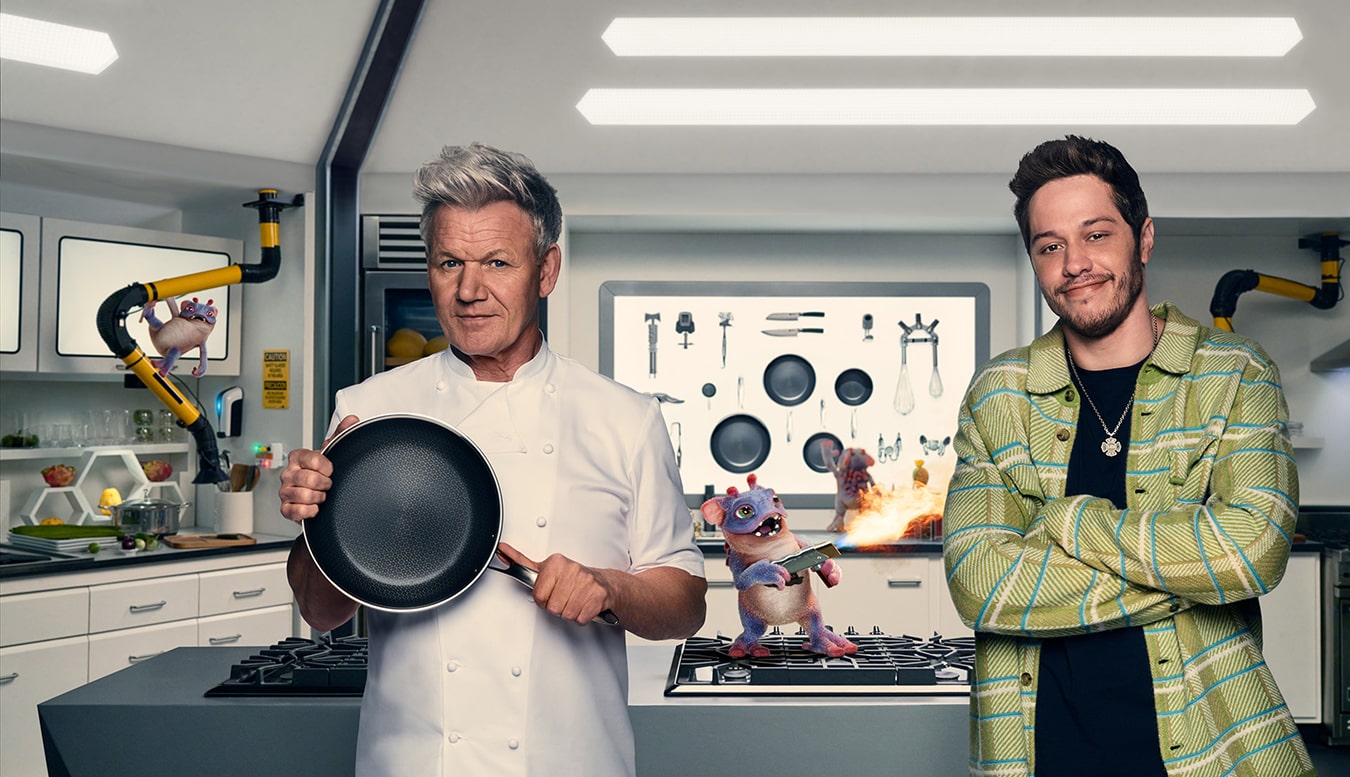 Two men in a kitchen with a cartoon creature holding a blowtorch.