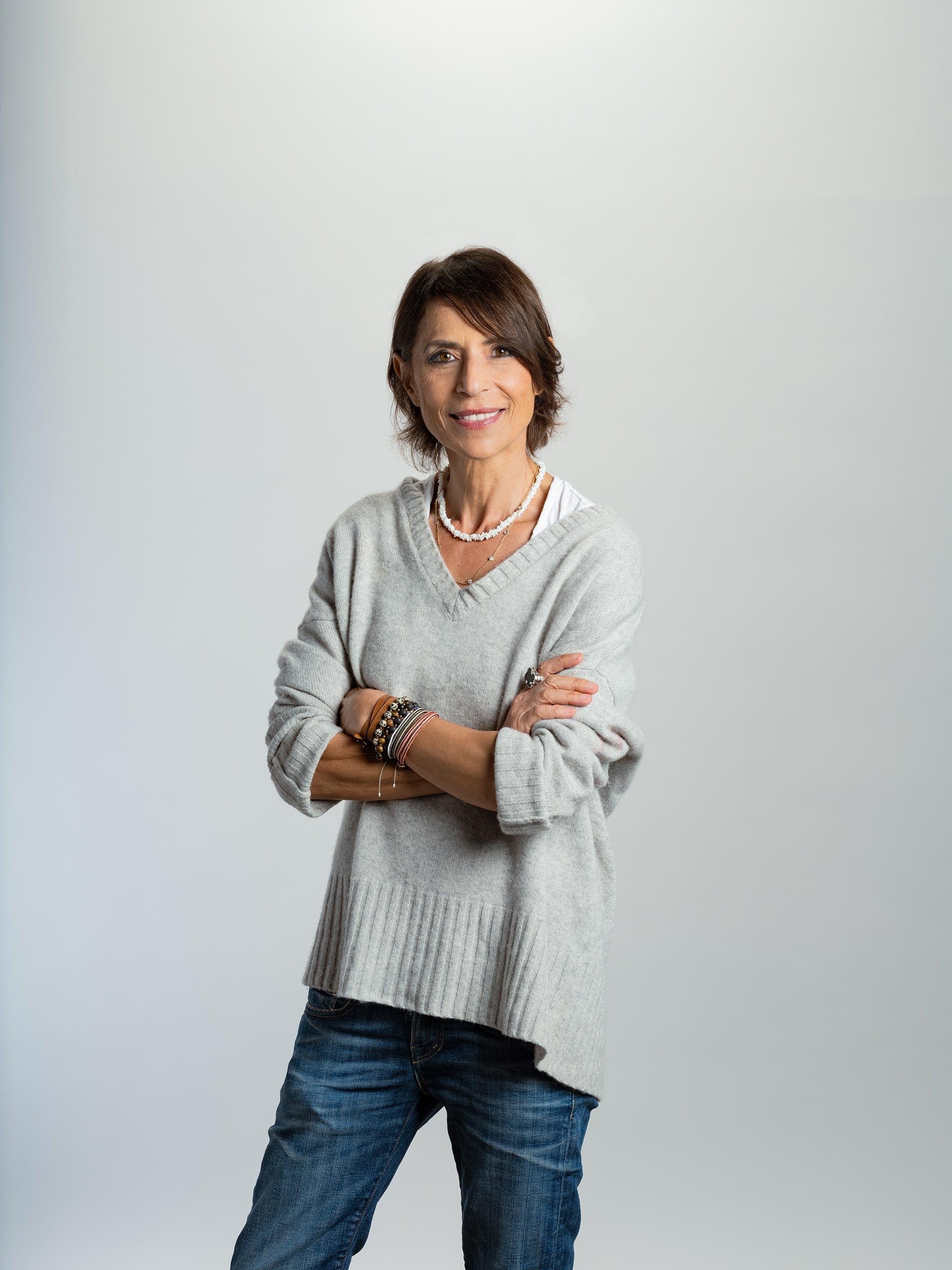 A person smiling, wearing a gray sweater and jeans, with crossed arms against a plain background.
