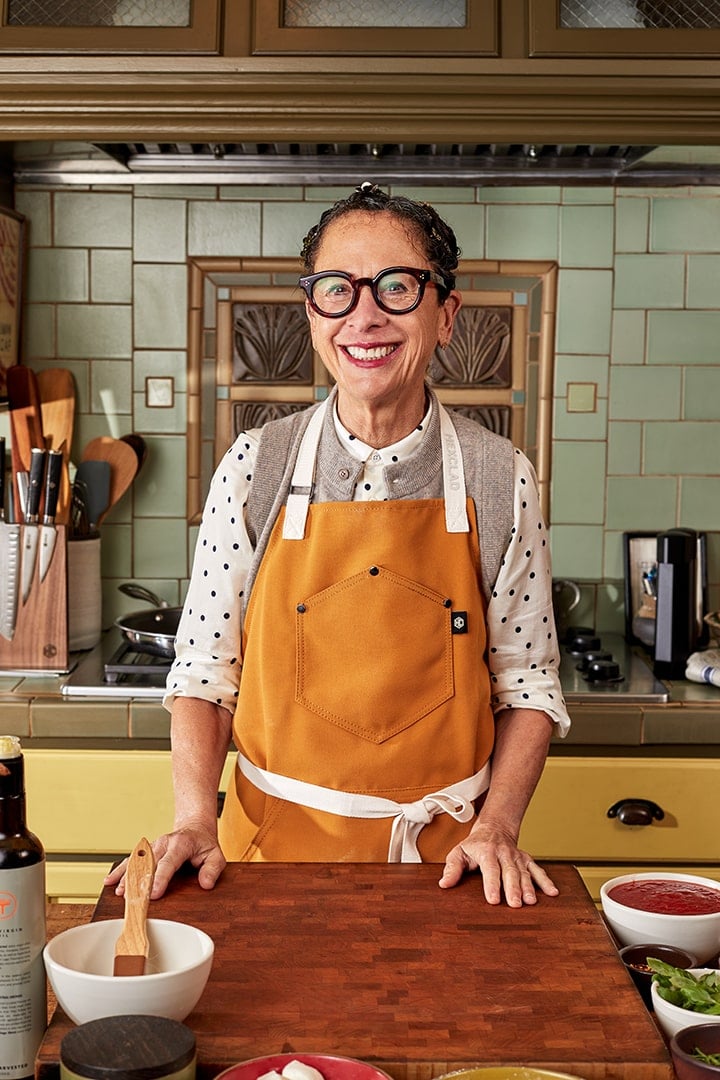 Smiling person wearing an apron stands in a kitchen with cooking utensils.