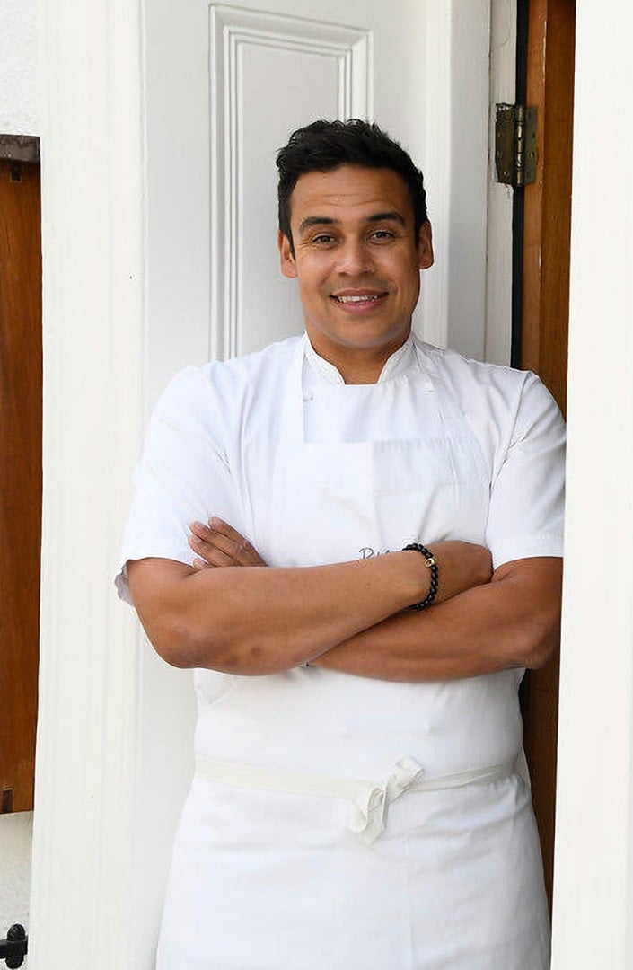 A chef wearing a white uniform stands smiling with arms crossed.