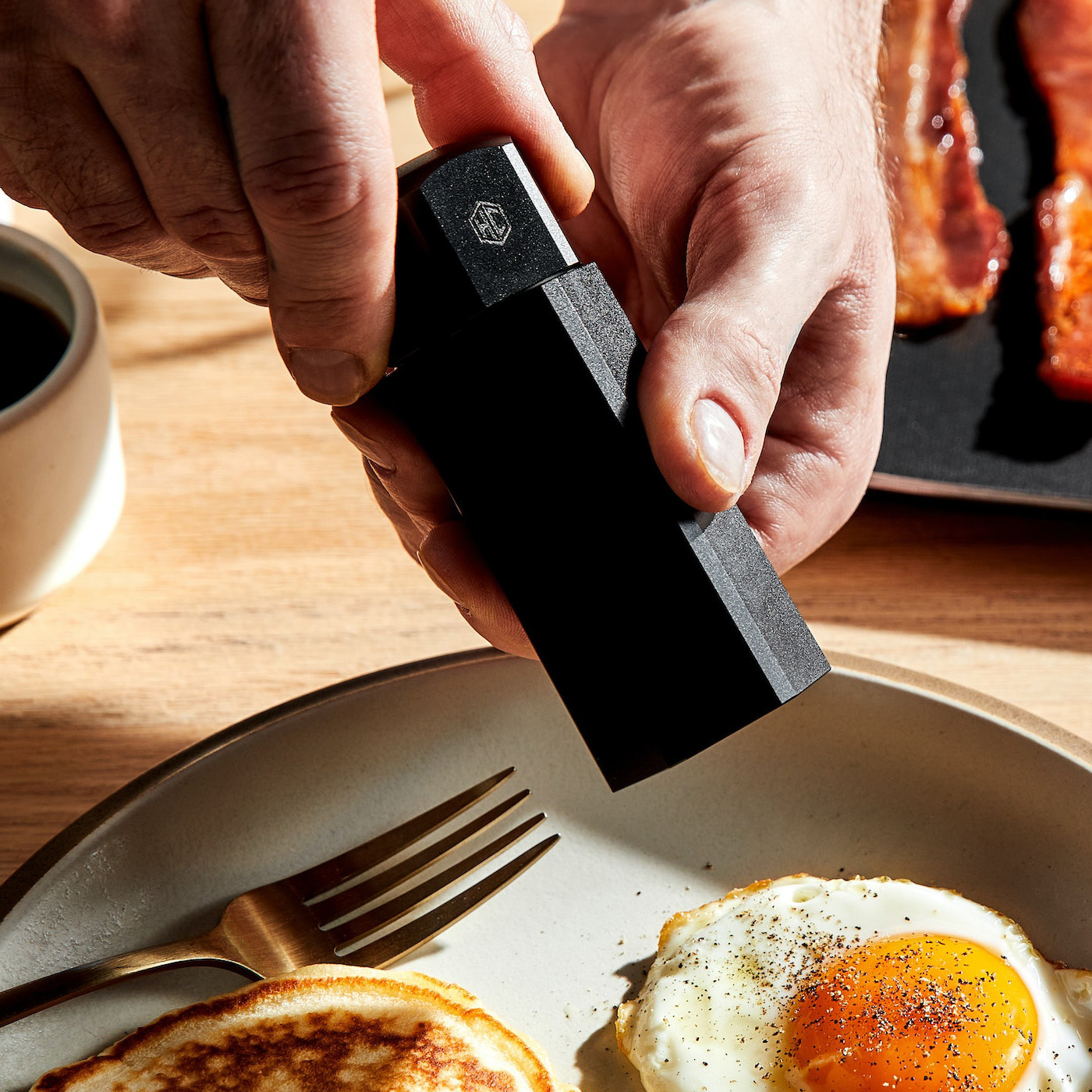 Close-up of a person seasoning an egg with a HexClad pepper grinder, with pancakes and bacon on the side, on a ceramic plate.