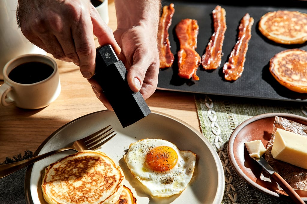 Eggs being peppered next to pancakes and bacon