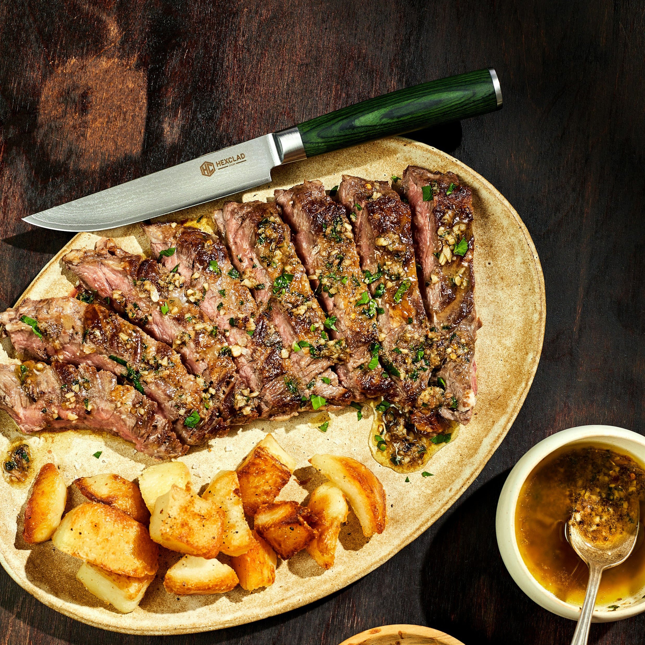 Sliced steak with garlic herb butter and roasted potatoes on a plate, accompanied by a knife and sauce.