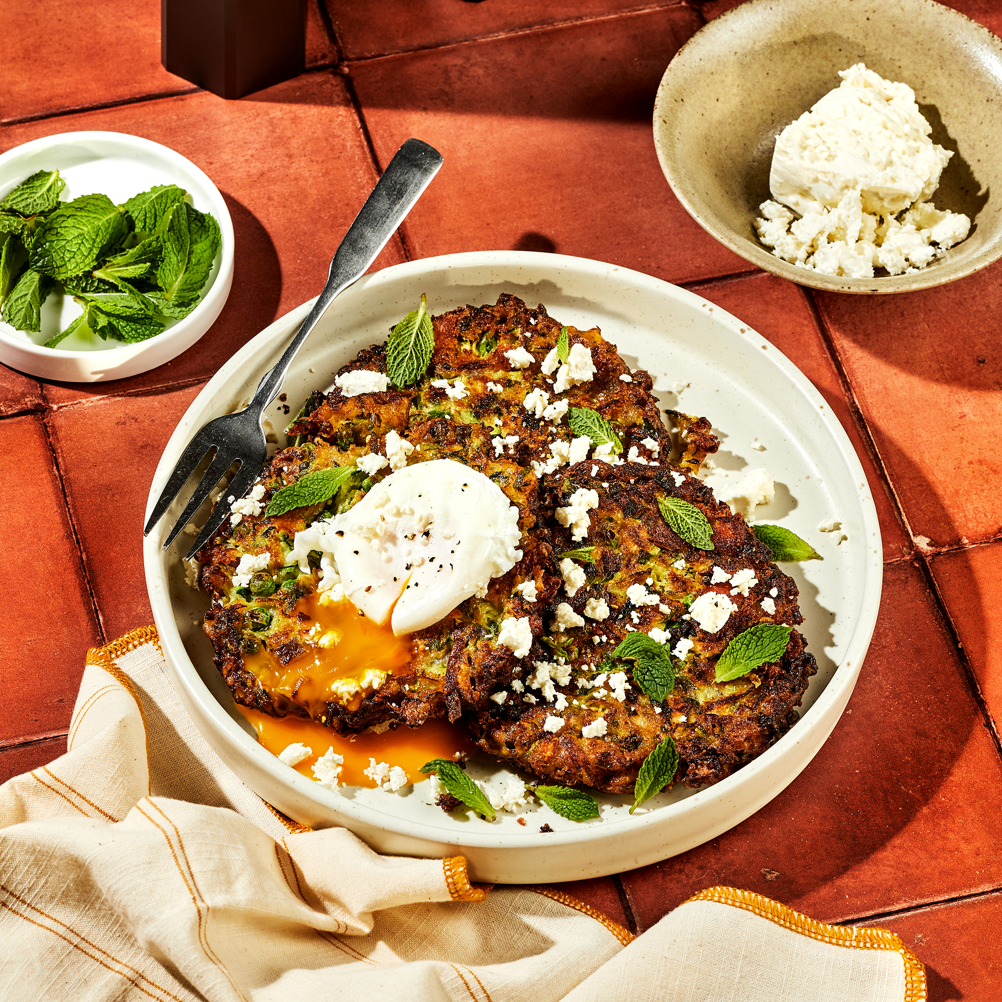 Plated fritters topped with crumbled cheese, mint leaves, and a runny poached egg.