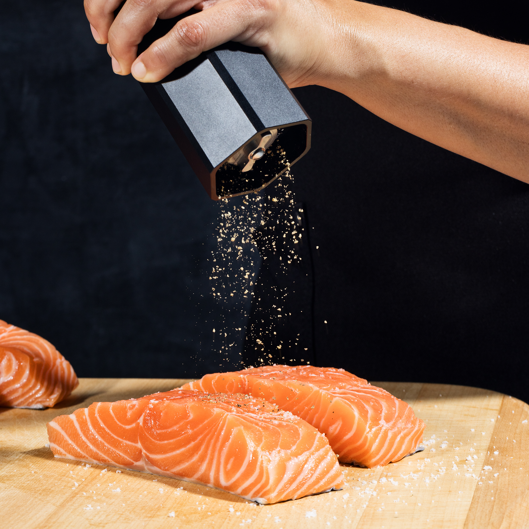 Hand seasoning fresh salmon fillets with black pepper on a wooden cutting board.