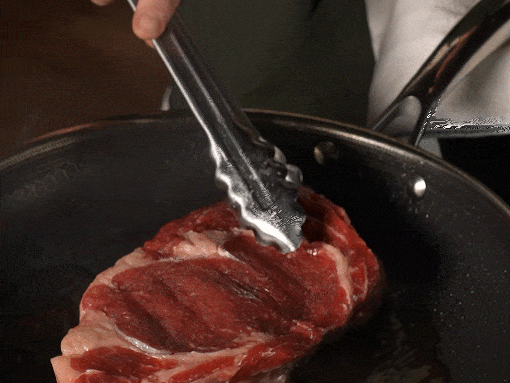 Person using tongs to place raw steak in a frying pan.