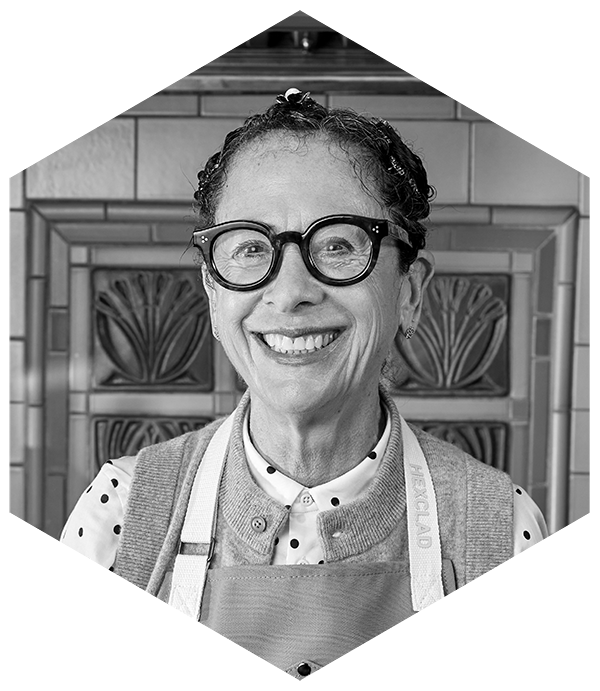 Smiling person with glasses, wearing a polka dot shirt and apron, standing in front of a decorative wall.