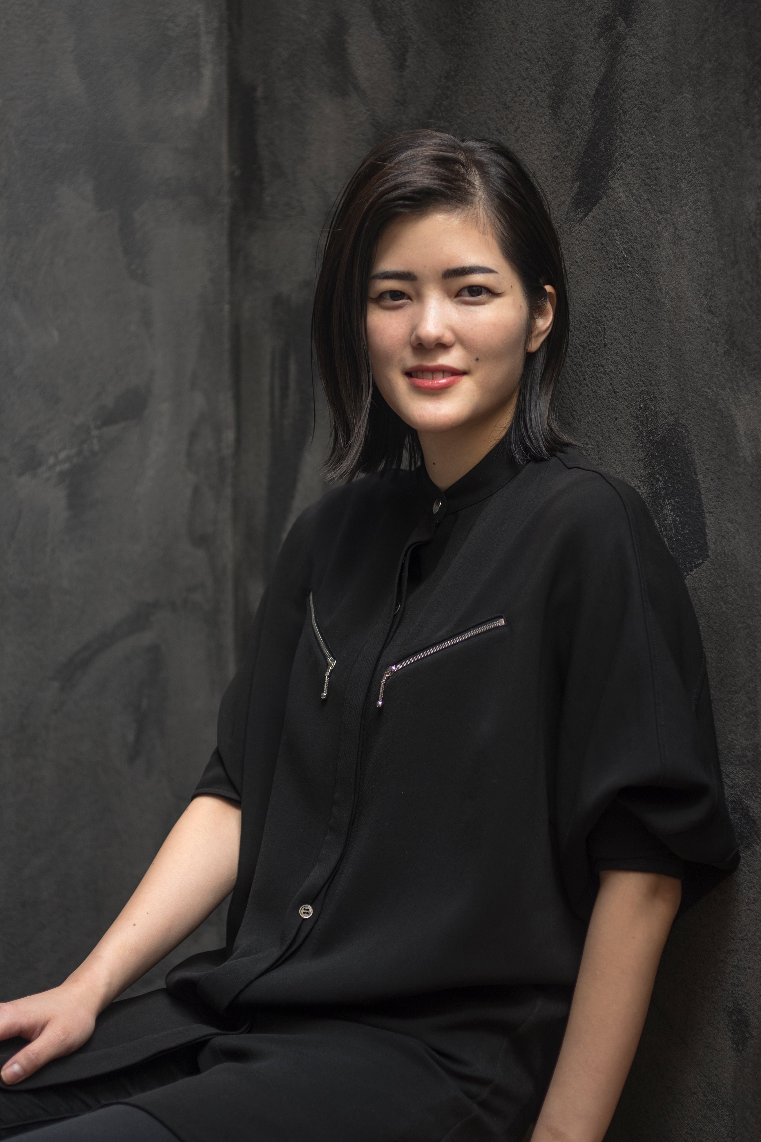 A person in a black outfit smiling against a dark textured background.