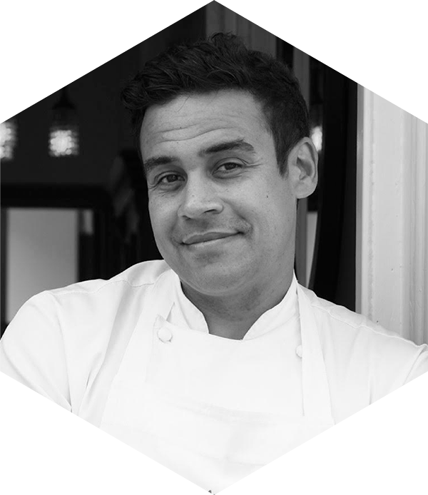 A chef smiling, wearing a white coat, in a black and white photo.