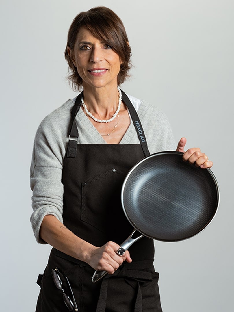 Woman in apron holding a frying pan against a neutral background.