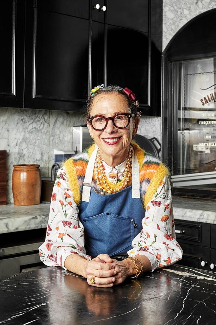 A person wearing glasses and colorful clothing smiling in a kitchen.
