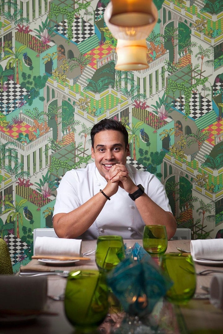 Chef smiling at a table with patterned wallpaper background.