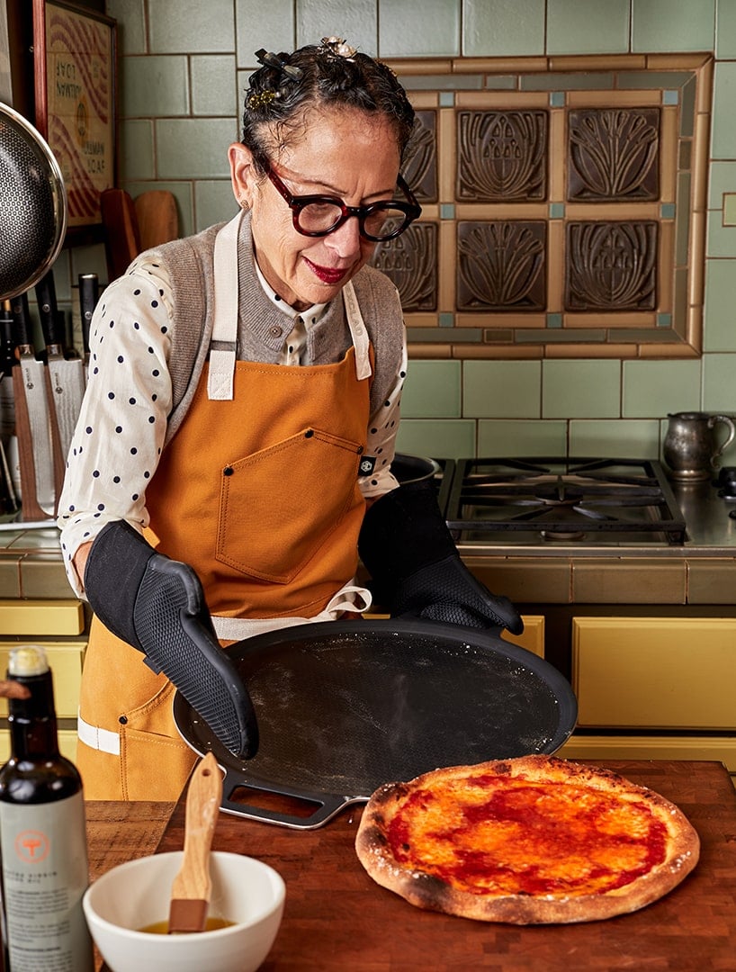 Person wearing an apron and gloves, holding a tray near a freshly baked pizza in a kitchen.