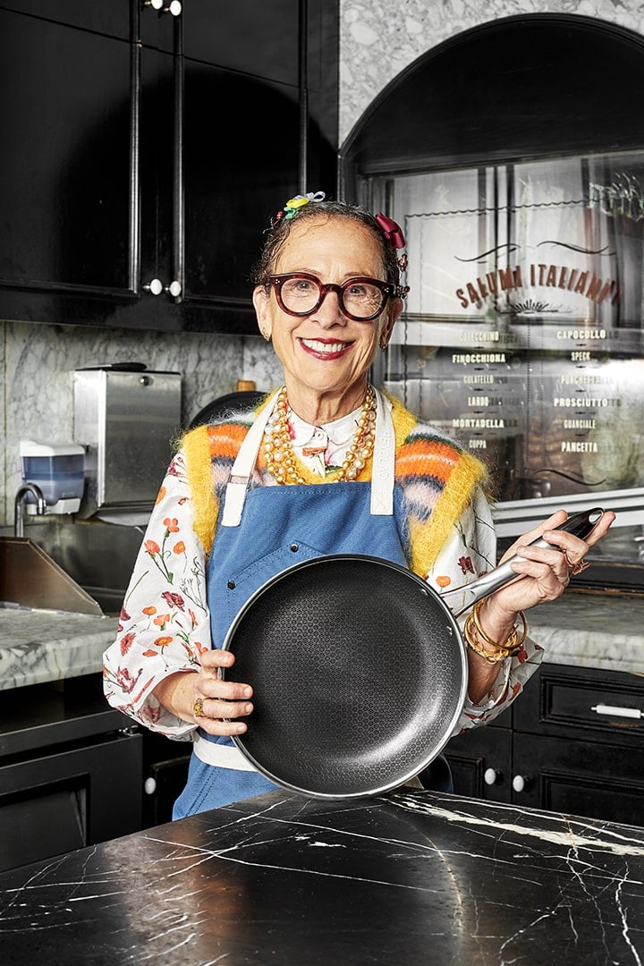 A smiling woman in a kitchen holds a frying pan, wearing a colorful outfit and an apron.