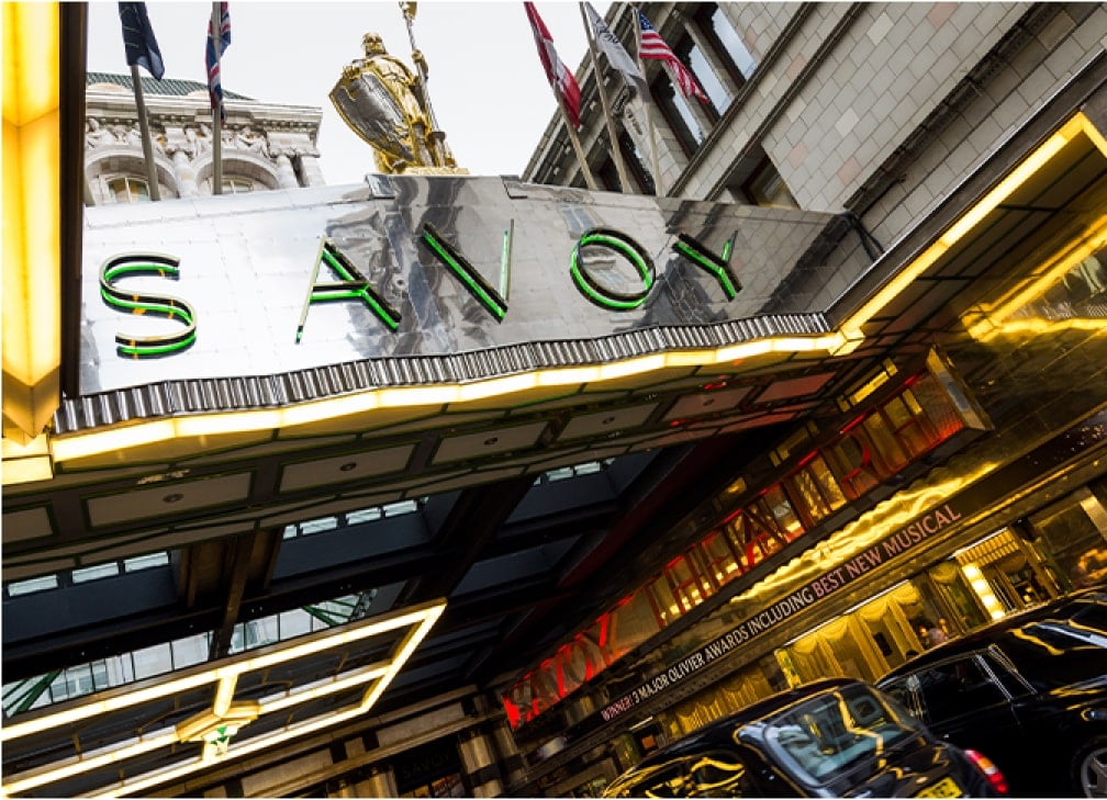 The exterior of The Savoy Hotel with illuminated signage and flags above the entrance.
