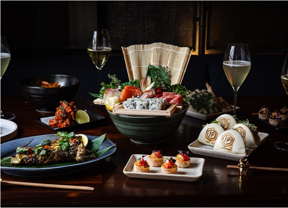 A table with various Japanese dishes, including sushi, baos, grilled fish, and wine glasses.