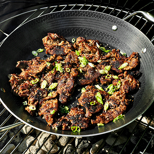 Grilled bulgogi (Korean marinated beef) in a pan on a barbecue grill, garnished with green onions.