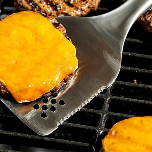 Burgers and buns grilling on a barbecue with a spatula in view.