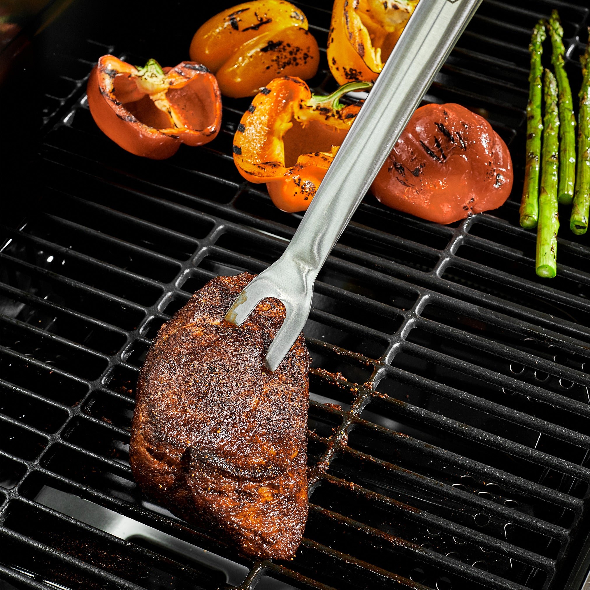 Grilled steak with tongs, alongside charred tomatoes and asparagus on a grill.