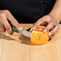 A person peeling an orange with a knife on a wooden surface.