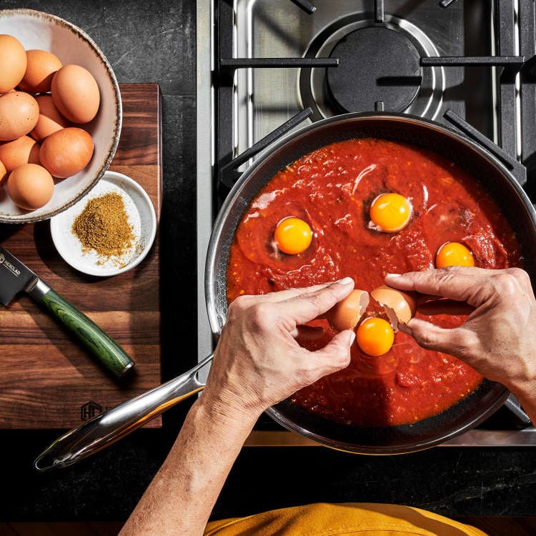 Person cracking eggs into a skillet of tomato sauce on a stove.
