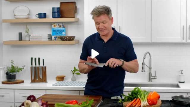 A person in a kitchen showing a knife with various vegetables on the counter.