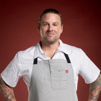 Chef wearing a white uniform and gray apron stands against a red background.