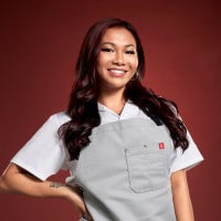 A smiling woman in a chef's uniform stands against a solid background.