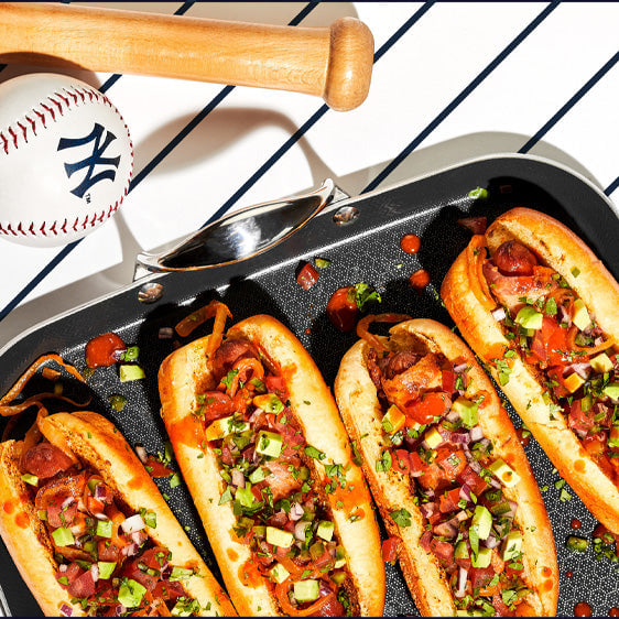 Hot dogs topped with vegetables in a grill pan, beside a baseball and bat.